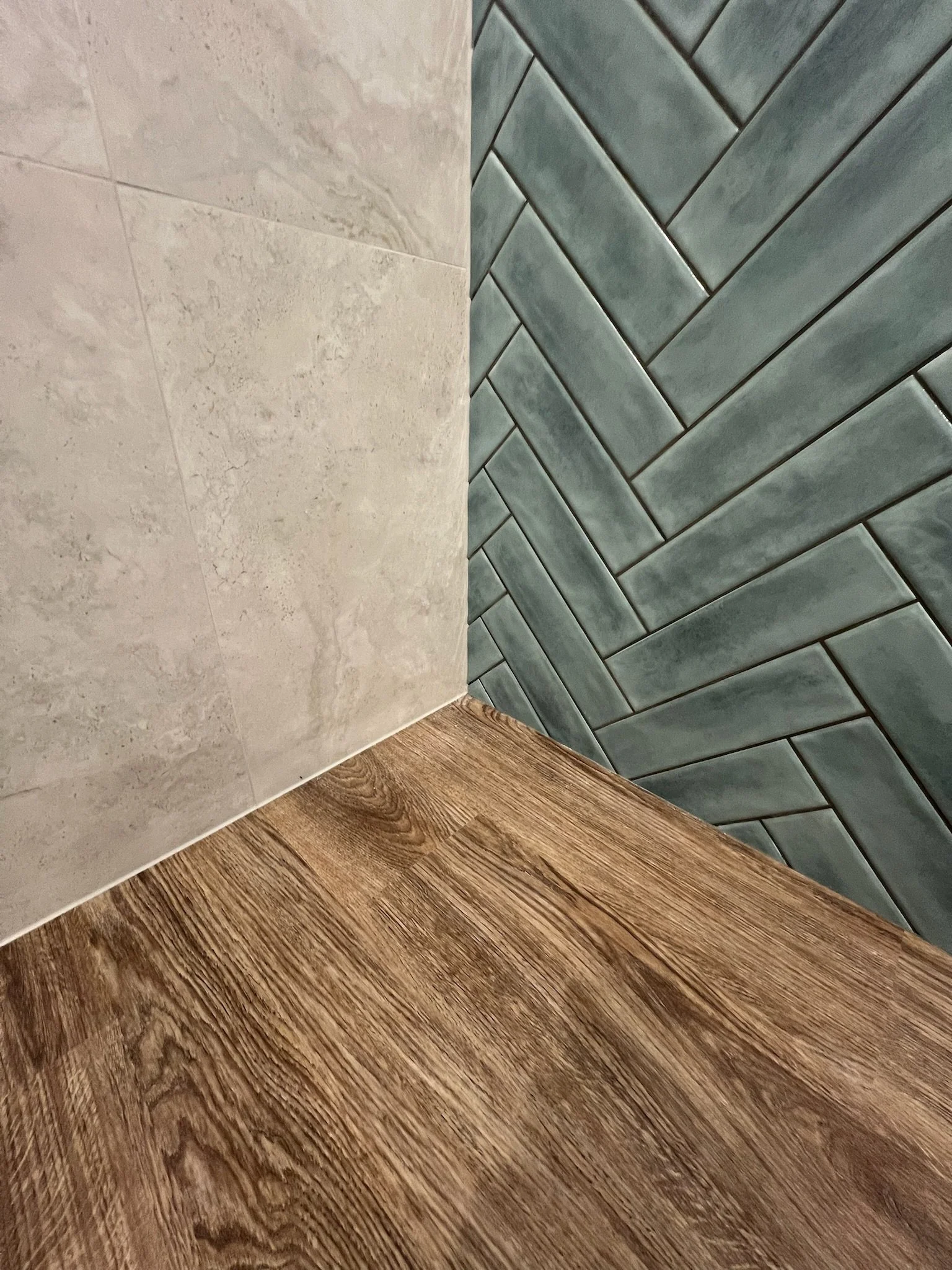 Interior corner of a room showing beige tiled wall, gray herringbone tiled wall, and wooden floor.