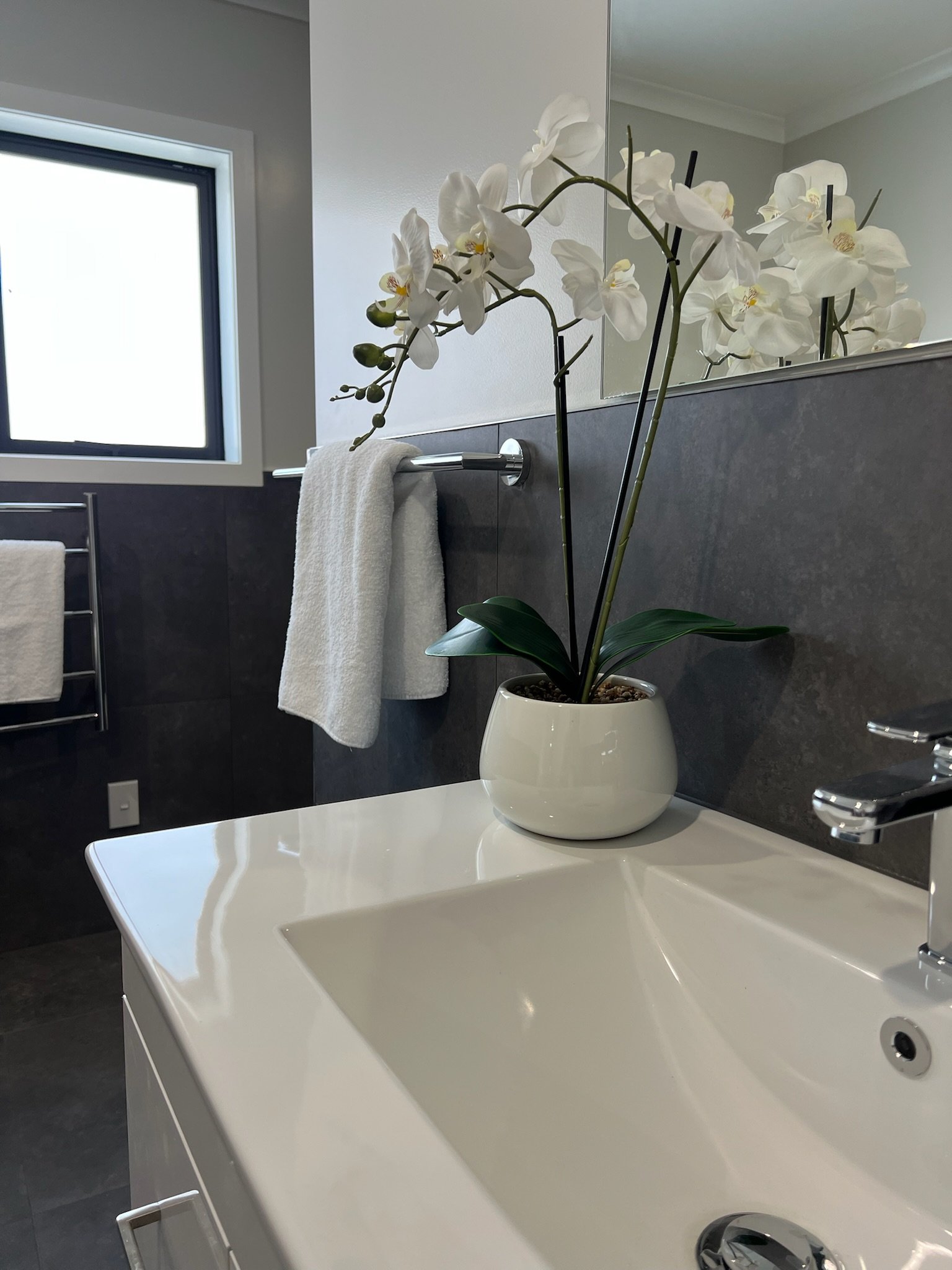 Bathroom sink with a white orchid in a white pot on the counter, towel rack with towels, window with frosted glass, mirror reflecting the orchid, gray tiles on the wall.