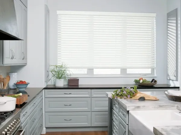 Bright kitchen with white cabinets, a window with blinds, a marble countertop, potted plant, bowls of tomatoes, and a cutting board with vegetables.