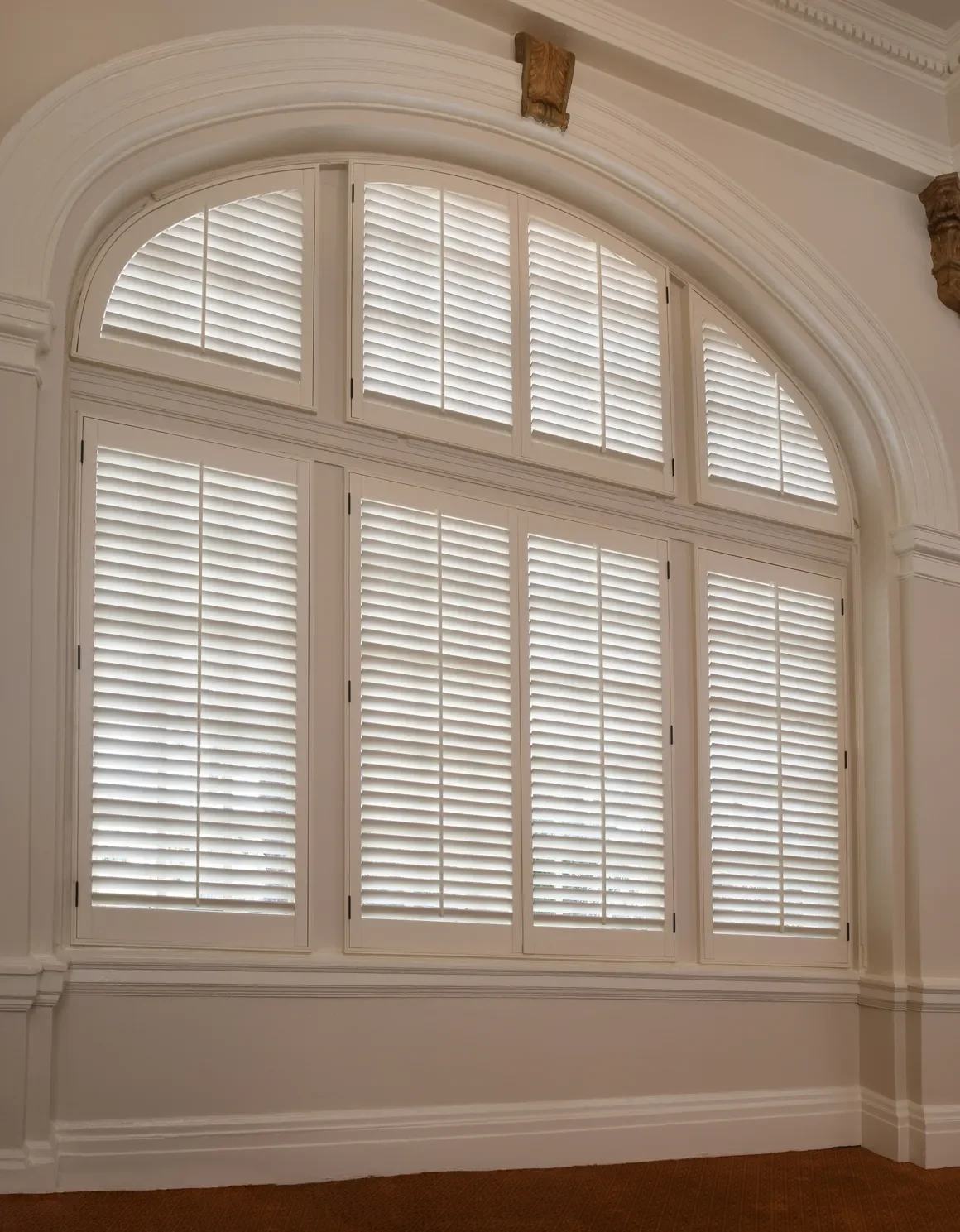 An elegant room with large arched windows with custom white wood shutters with traditional front tilt rods, arch top shutters.