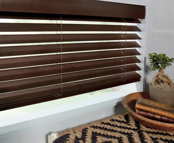 Close-up of a window with dark brown horizontal blinds, partially open, with a potted plant and a woven basket with rolled-up fabric or towels nearby.
