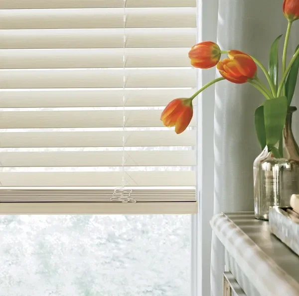 A window with beige horizontal blinds partially open, with a glass vase of orange and yellow tulips placed on a white surface nearby.