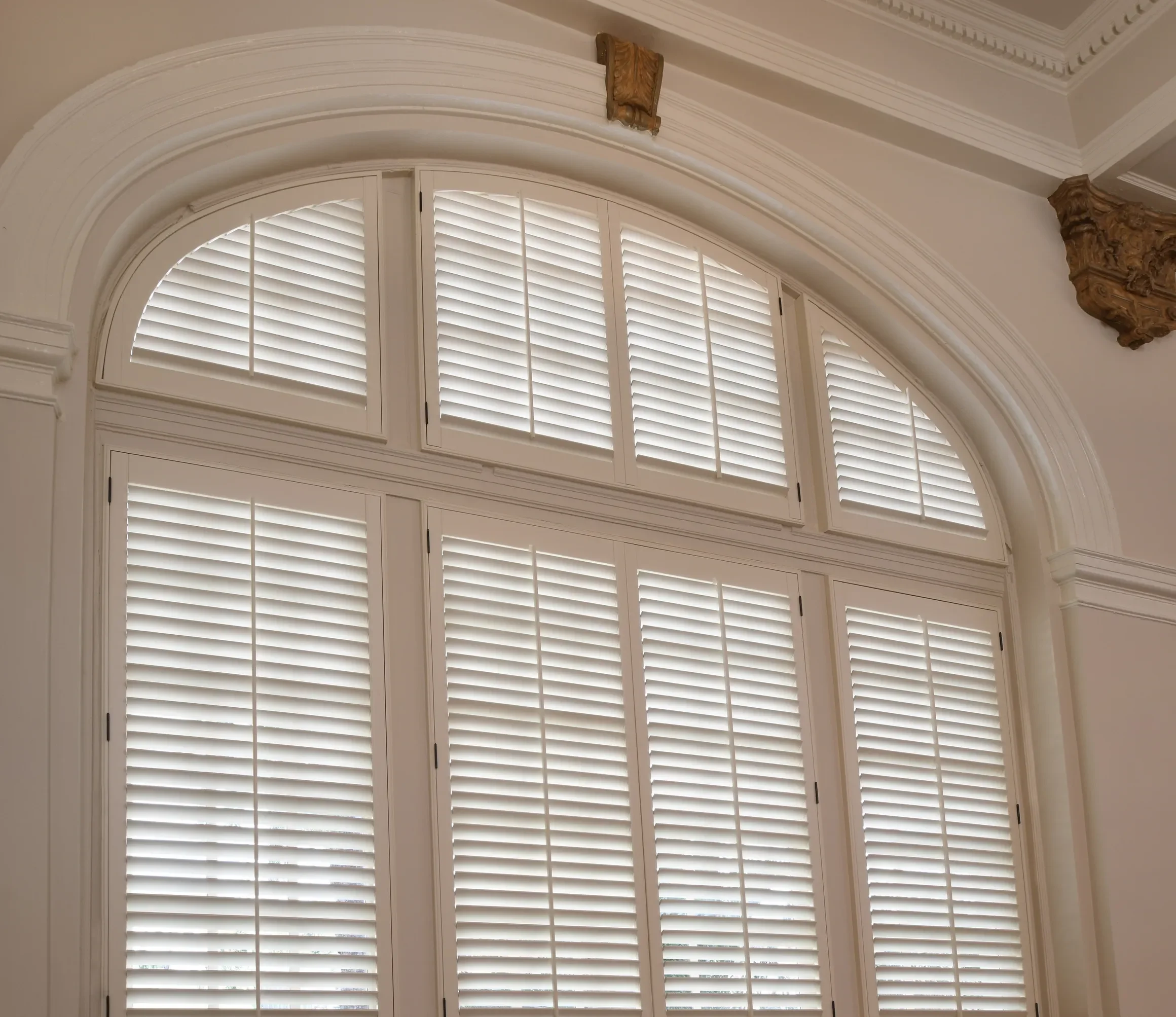 An elegant room with large arched windows with custom white wood shutters with traditional front tilt rods, arch top shutters.