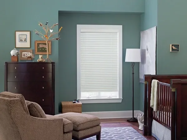 A cozy nursery room with teal walls, a window with blinds, a dark wood dresser with decorative items on top, a beige armchair, a wicker basket, and a crib with a towel hanging over the side. There's also a floor lamp, framed pictures, and a large abs