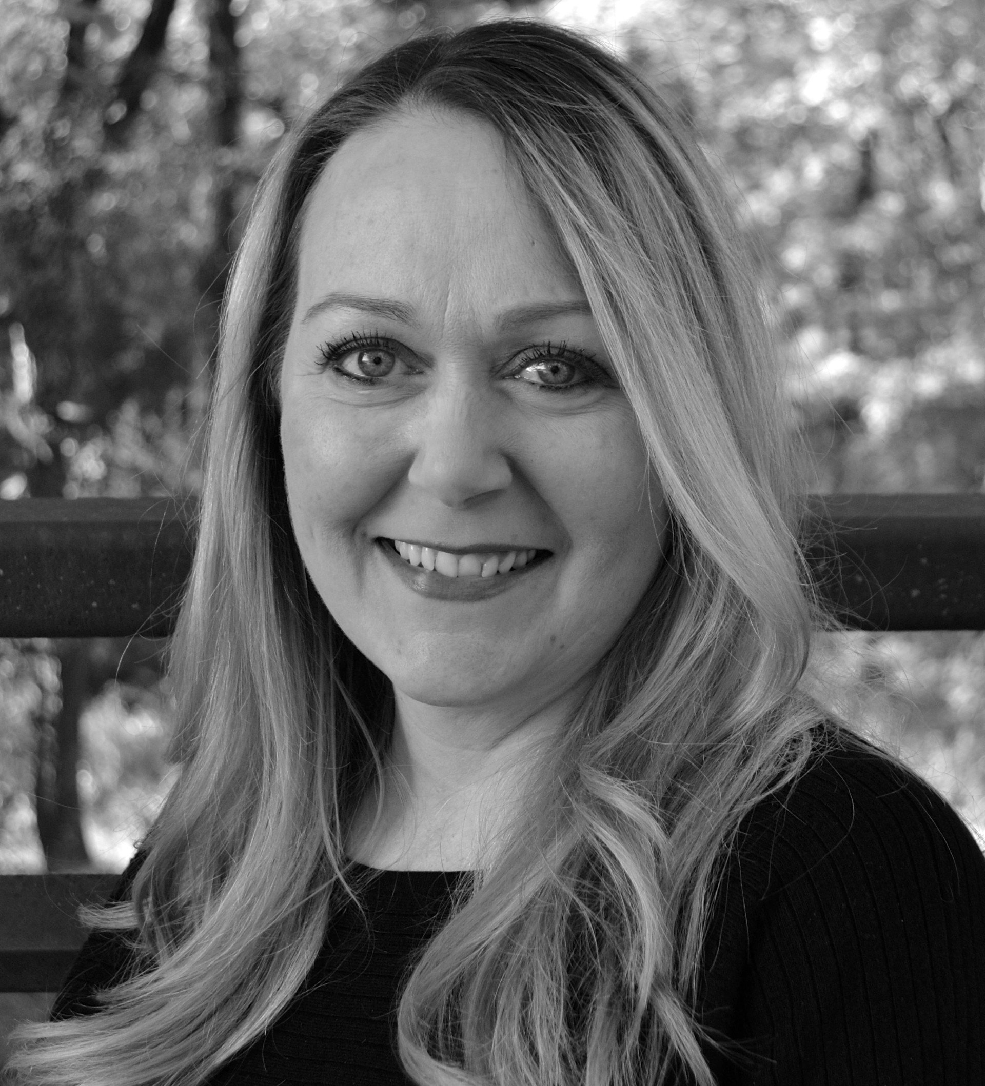 A black and white photograph of a woman with long wavy hair, smiling, wearing a dark top, outdoors with trees in the background.
