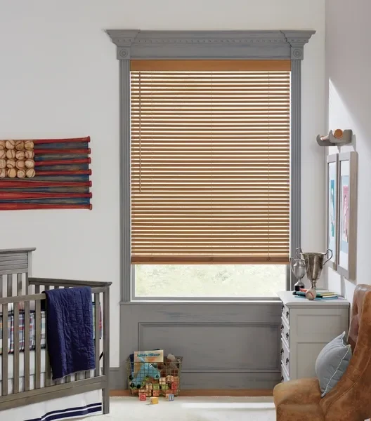 A nursery room with a window covered by wooden blinds, gray decorative trim, a crib with a navy blanket, a small white dresser with trophies and books, and framed artwork on the wall.