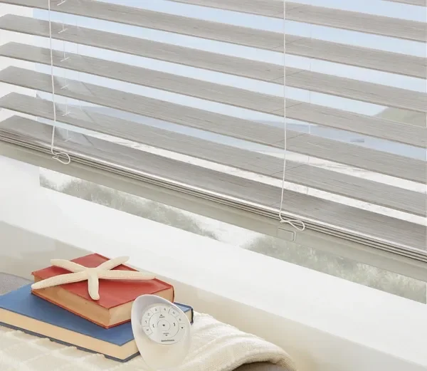 Close-up of an interior window with closed blinds, with a white alarm clock and a small unidentifiable object on top of a stack of books, all on a windowsill.