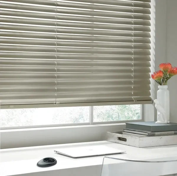 A window with beige horizontal blinds, a white desk with a closed laptop, a black smart device, gray and white books, and a white vase with orange flowers.
