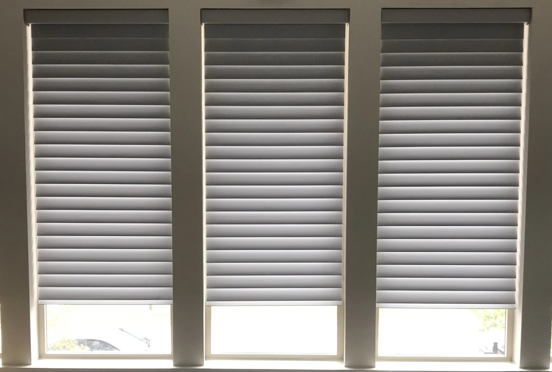 Three windows with white horizontal blinds, partially open, with outside view showing a car and some greenery.