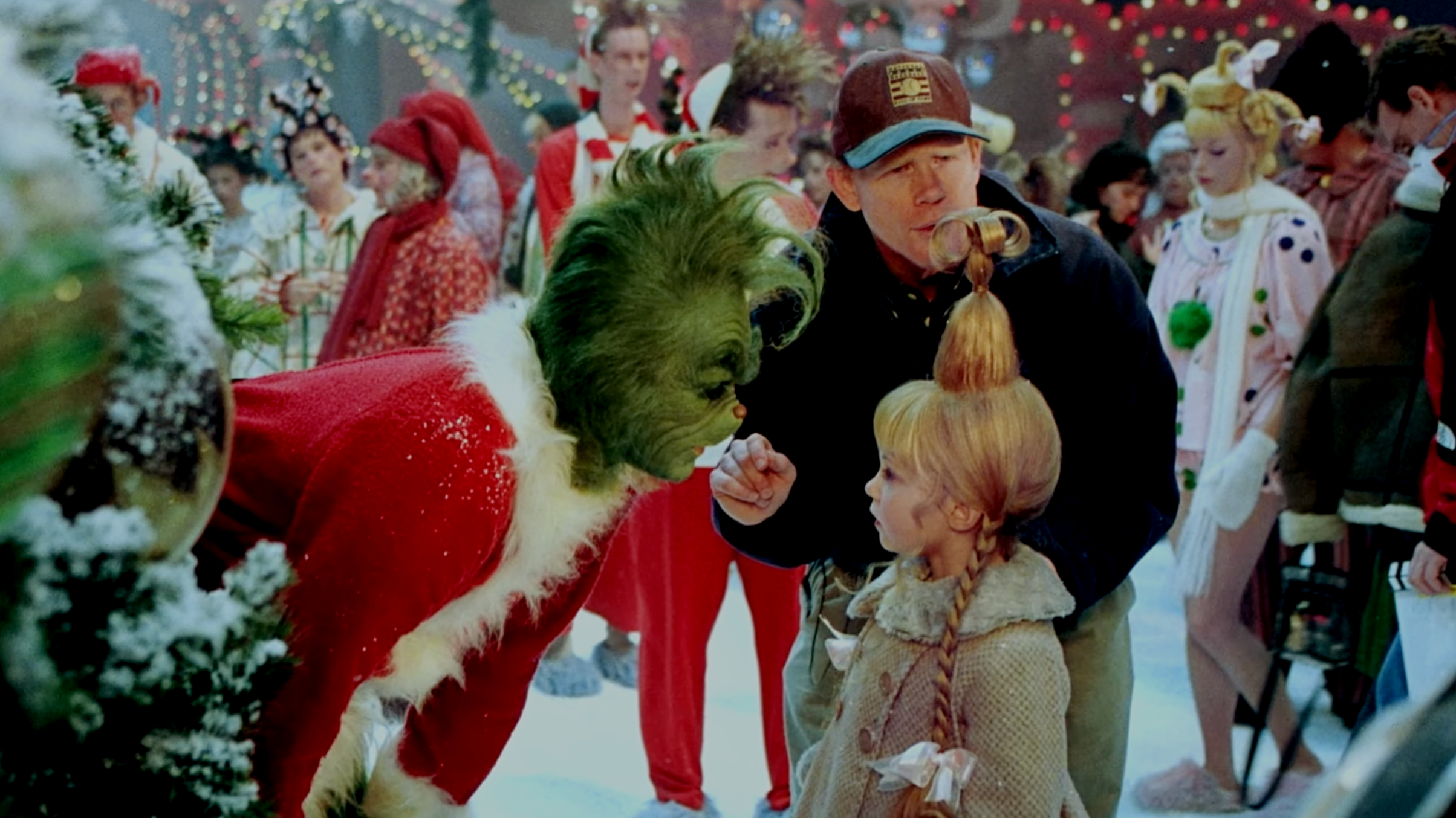A green-faced creature dressed as Santa Claus is leaning toward a little girl with red hair and a braided hairstyle, who is standing with her arms by her side. An adult man with a cap and jacket is touching the girl's hair. The scene is set at a lively, decorated Christmas party with people in festive costumes and colorful lights.