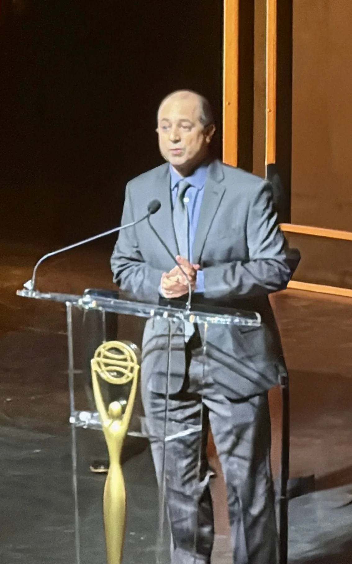 A man in a gray suit giving a speech at a podium with a large gold trophy on it.