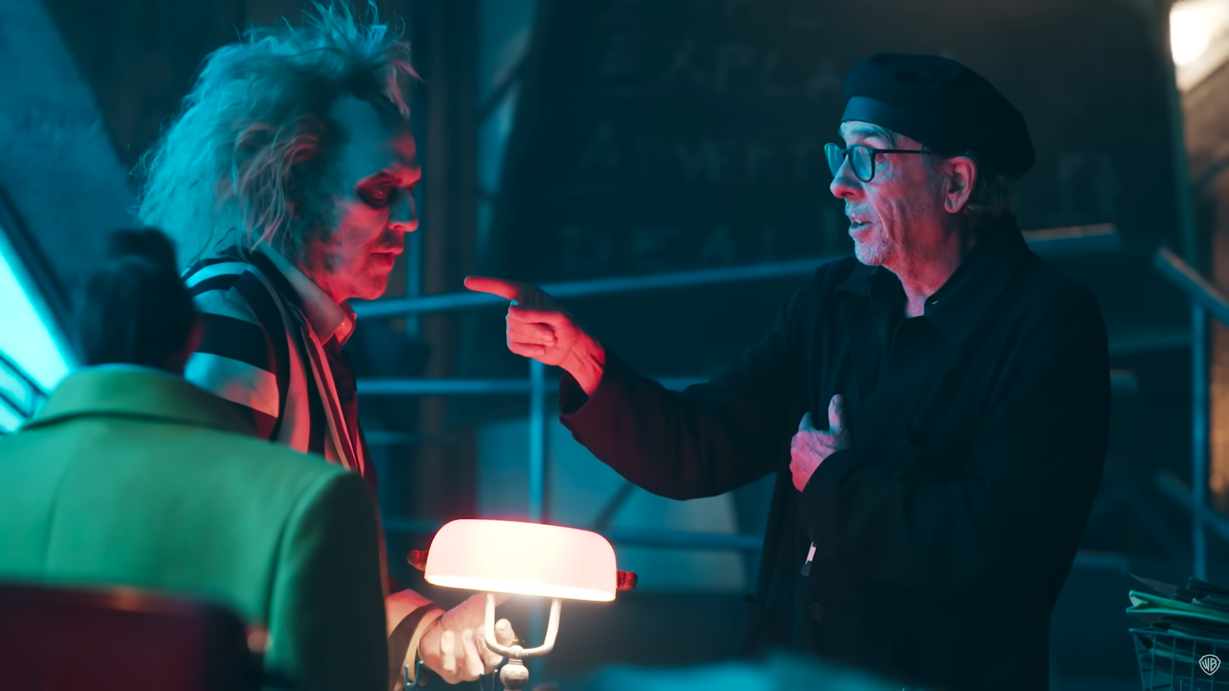 A woman with light blond hair and heavy makeup, wearing a black and white striped jacket, is holding a small lamp and looking down. An older man with glasses, a black beret, and a black shirt is pointing at her with an intense expression. The scene has moody, colorful lighting.