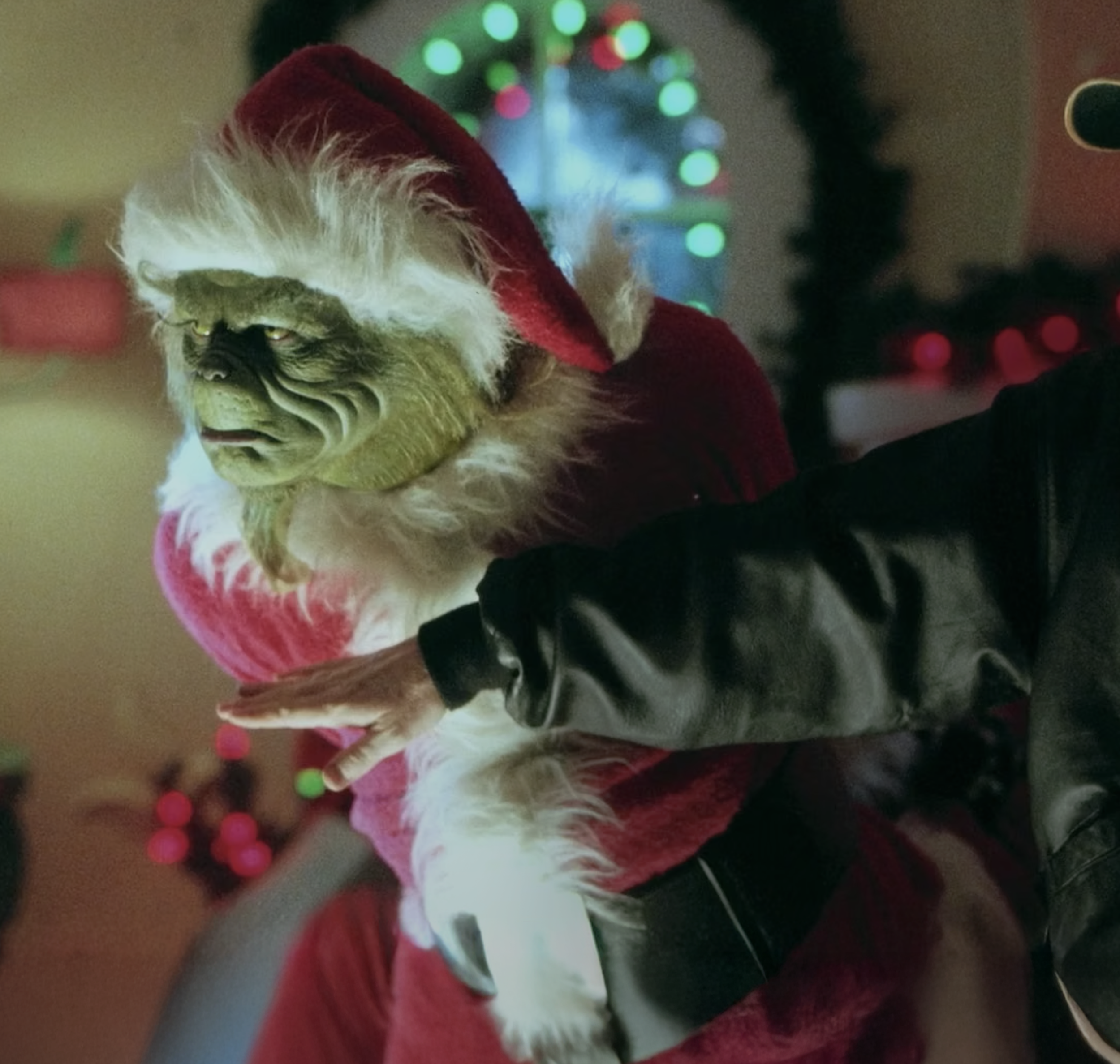 A green-faced creature dressed as Santa Claus is leaning toward a little girl with red hair and a braided hairstyle, who is standing with her arms by her side. An adult man with a cap and jacket is touching the girl's hair. The scene is set at a lively, decorated Christmas party with people in festive costumes and colorful lights.