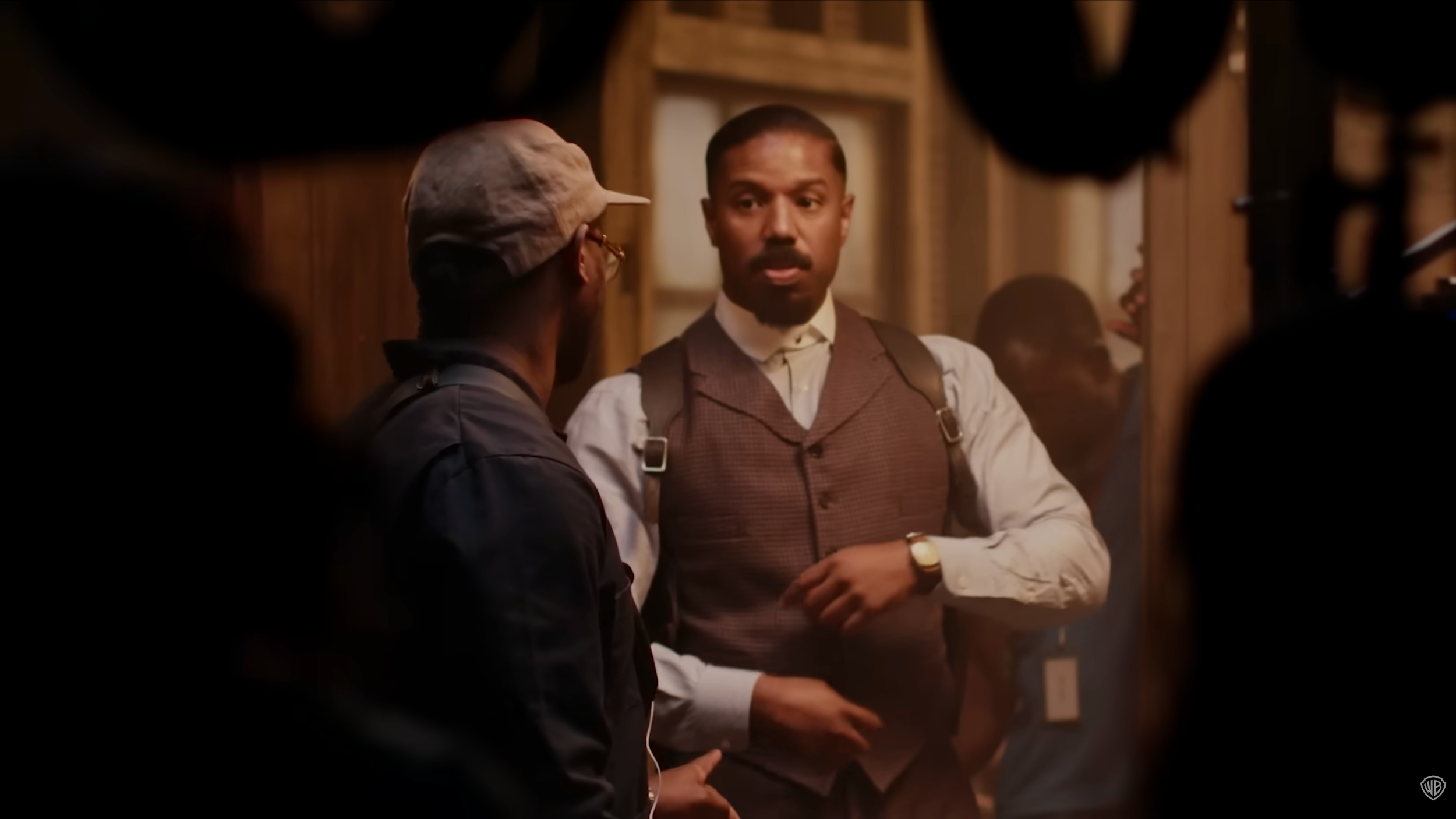 Two men in a wood-paneled room engaged in conversation, one wearing a hat and glasses, the other with a mustache and suspenders, looking at each other in a scene from a period film.