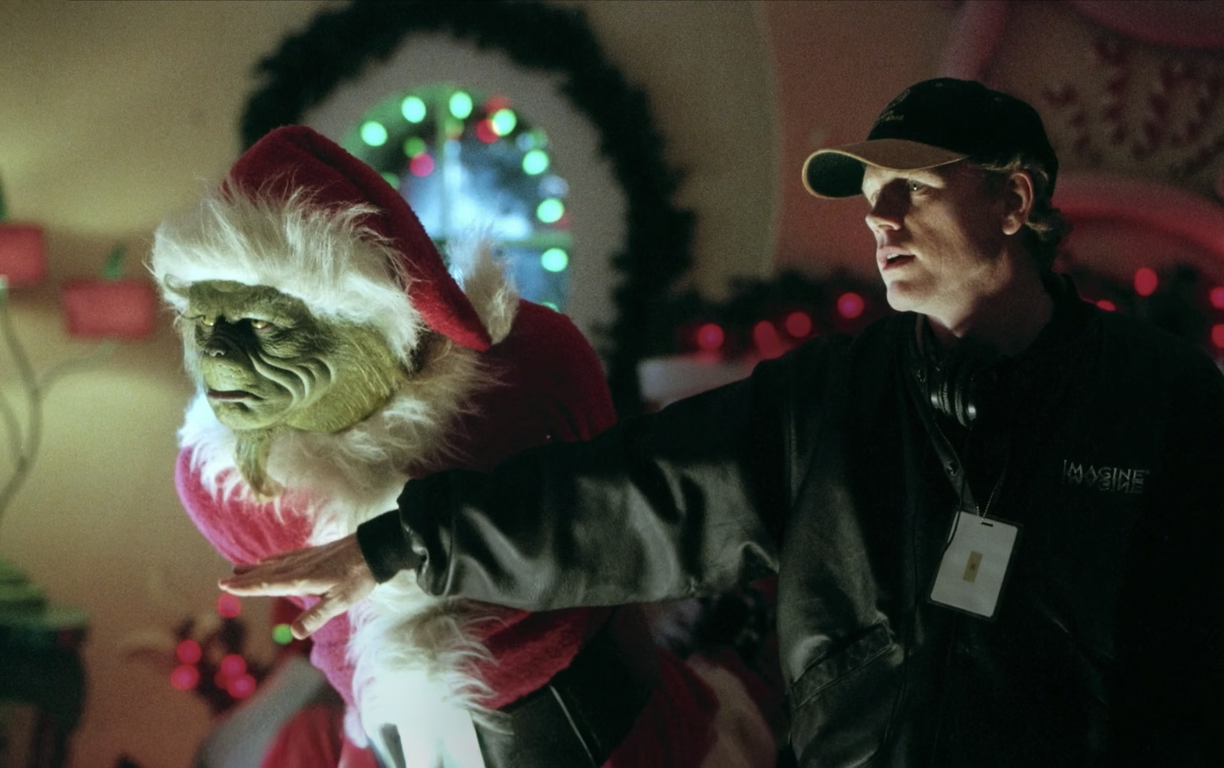 A green-faced creature dressed as Santa Claus is leaning toward a little girl with red hair and a braided hairstyle, who is standing with her arms by her side. An adult man with a cap and jacket is touching the girl's hair. The scene is set at a lively, decorated Christmas party with people in festive costumes and colorful lights.