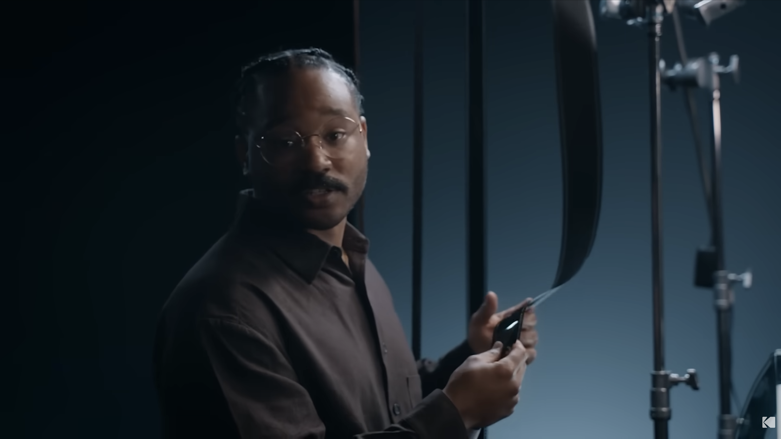 A man with glasses and a mustache holds a filmstrip and presents it to the camera, with film equipment visible in the background.