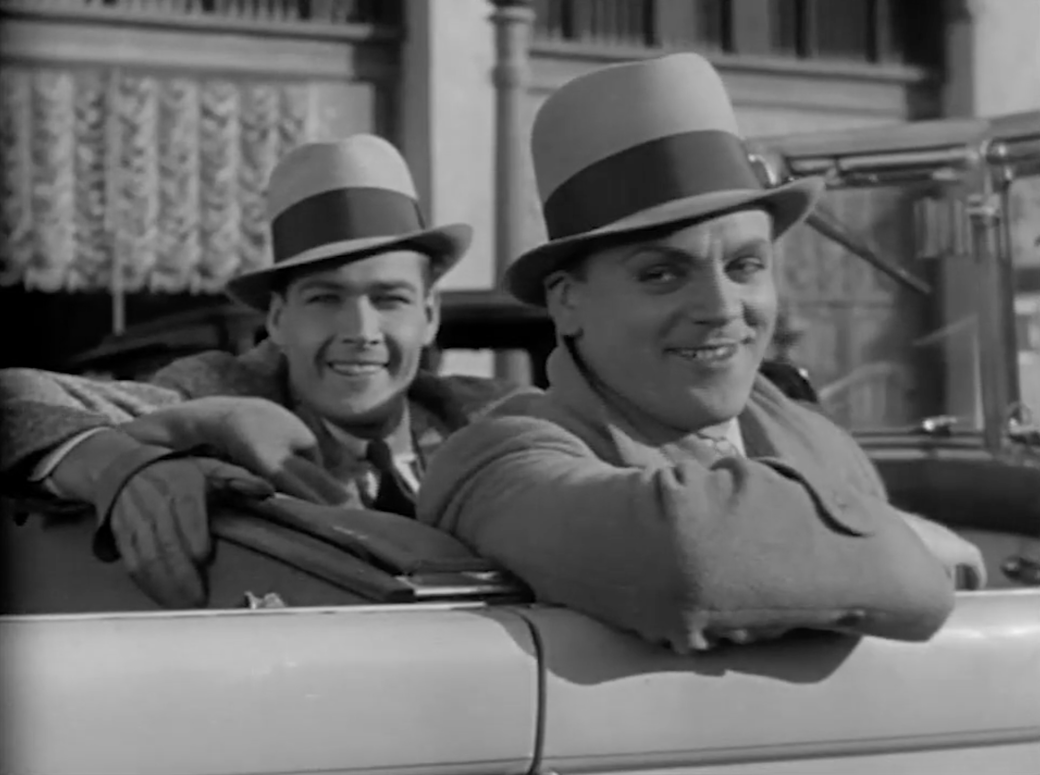 Two men smiling and leaning on a car's window, both wearing hats and coats, with a background that appears to be a street scene.