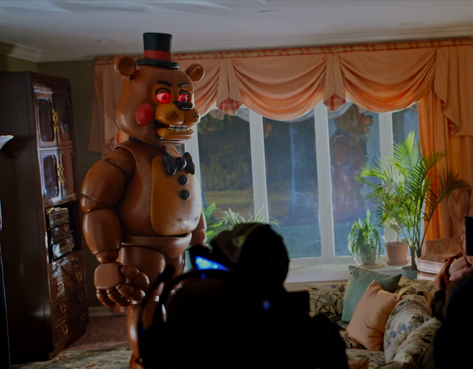 A person filming a person in a bear costume with glowing red eyes in a living room.