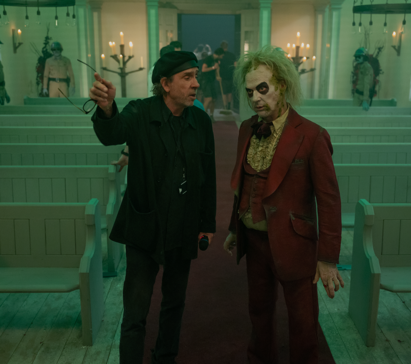 A woman with light blond hair and heavy makeup, wearing a black and white striped jacket, is holding a small lamp and looking down. An older man with glasses, a black beret, and a black shirt is pointing at her with an intense expression. The scene has moody, colorful lighting.