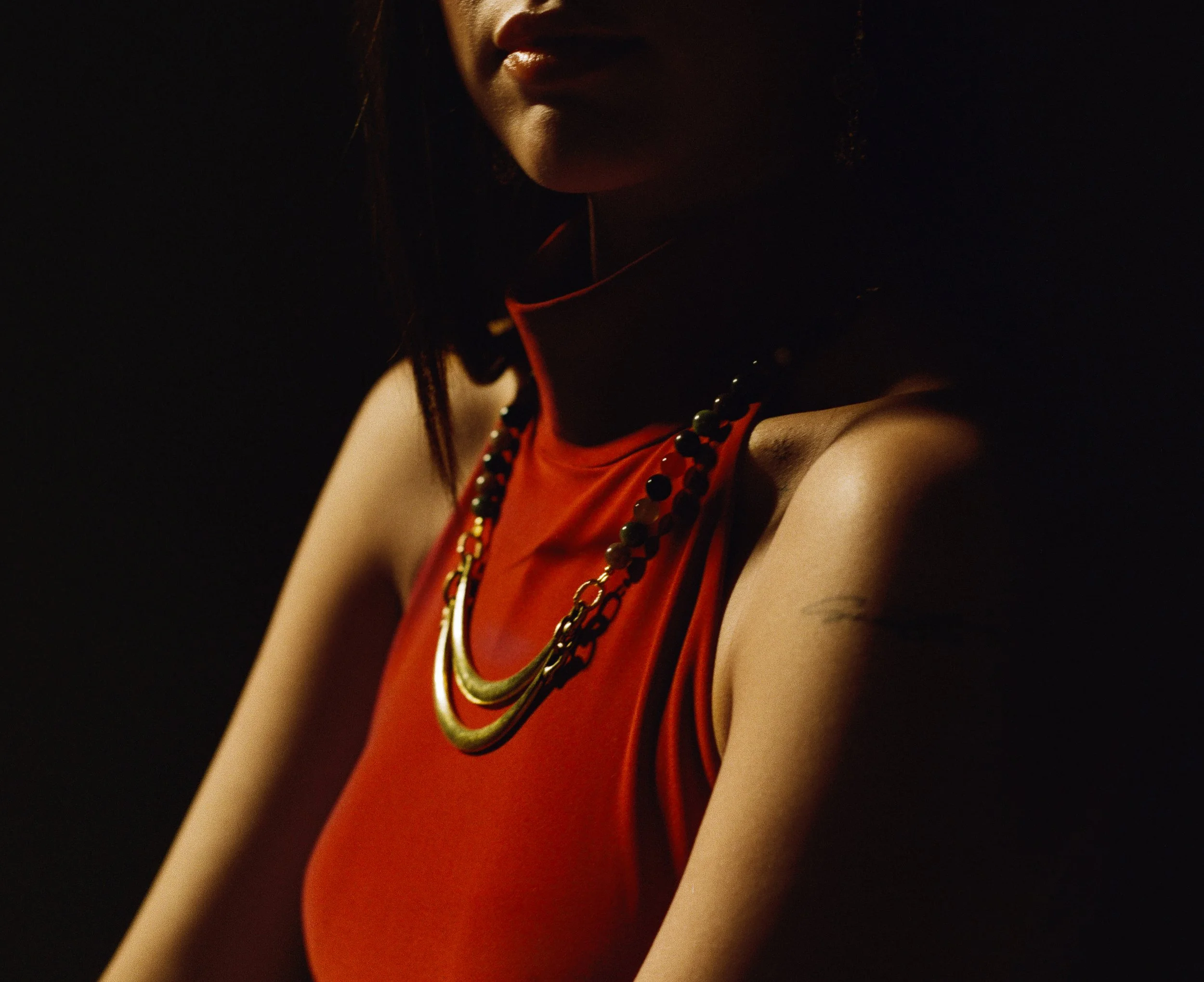 Woman wearing a red sleeveless top and layered necklaces, with part of her face visible against a dark background.