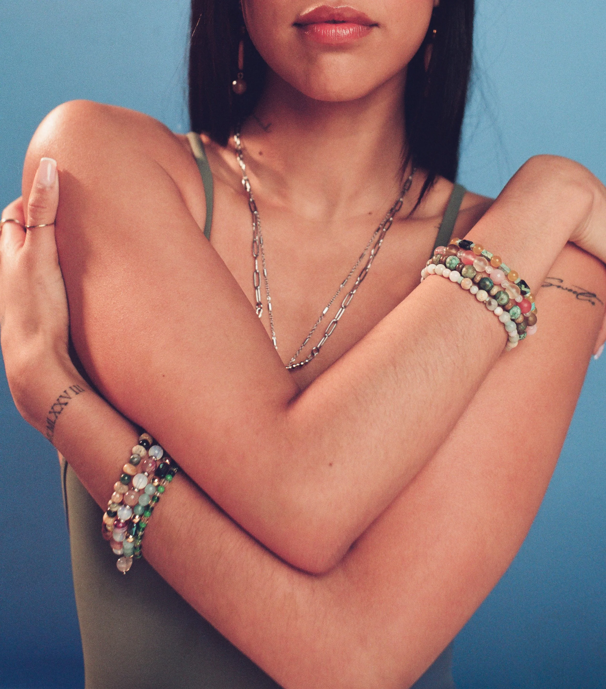 A woman with dark hair and multiple beaded bracelets on her arms, wearing layered necklaces, a green tank top, and gold earrings, standing against a blue background.