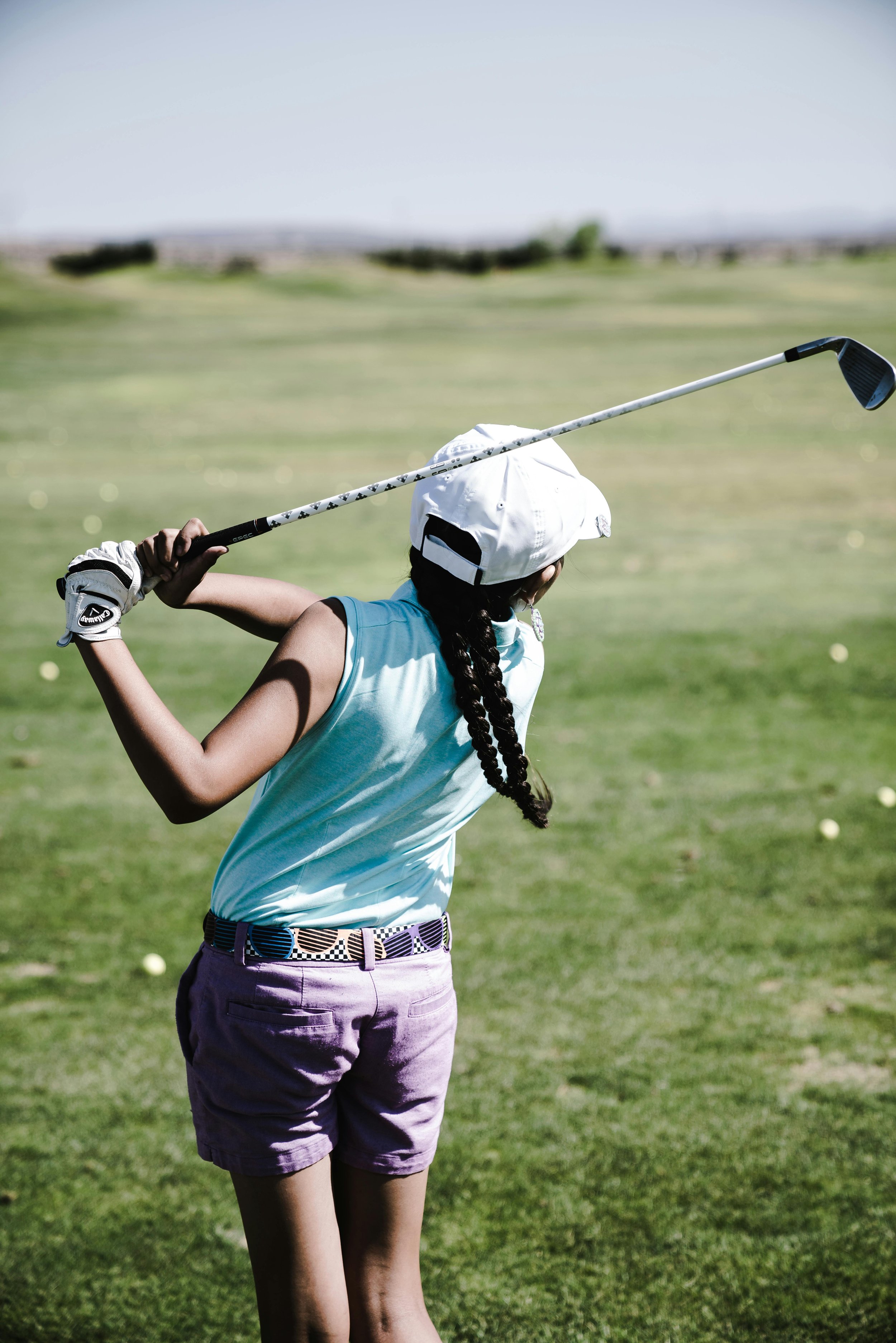 A girl wearing a white cap, sleeveless turquoise top, and purple shorts practicing golf on a sunny golf course, holding a golf club over her shoulder.