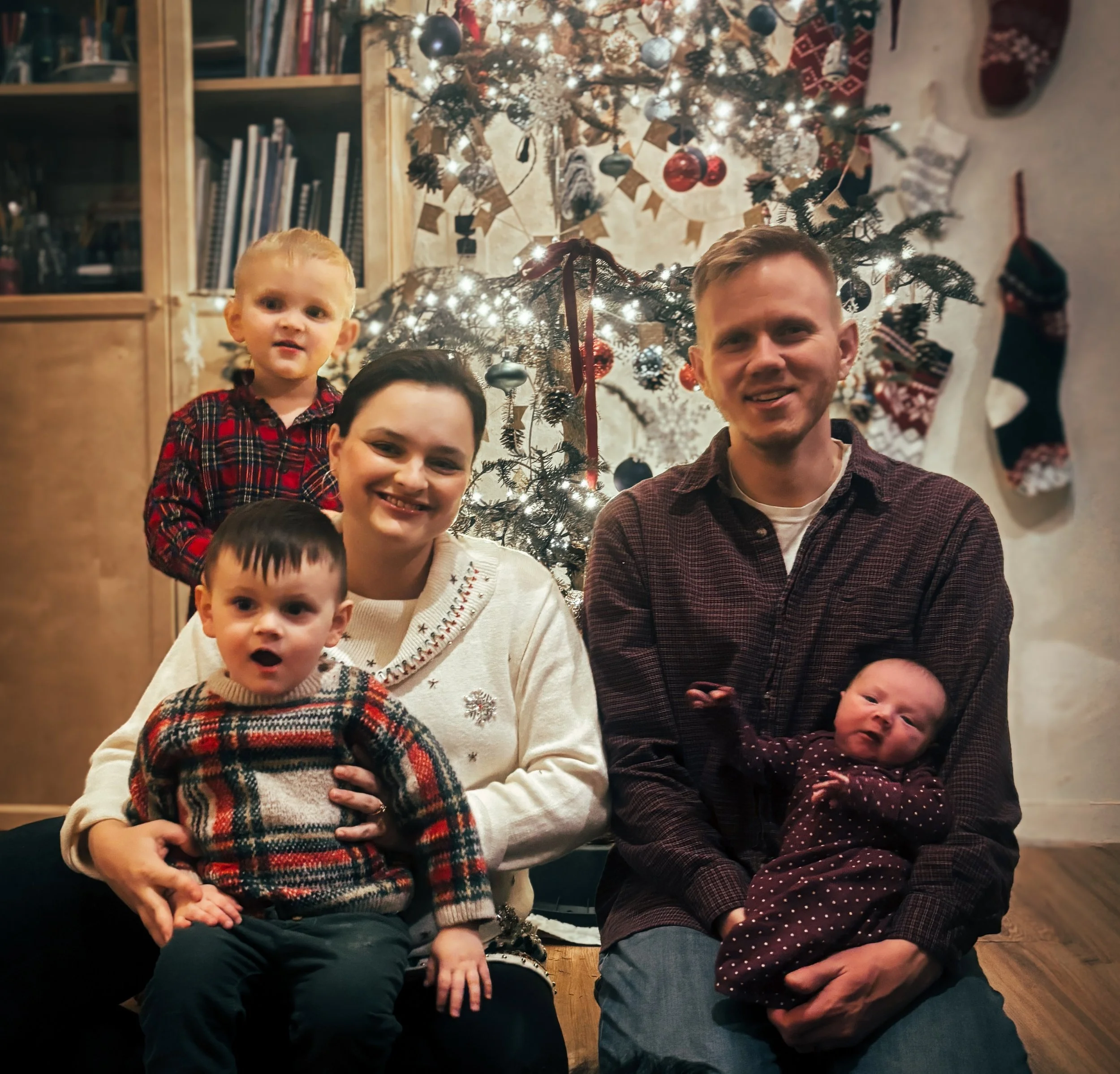 A family of five celebrating Christmas in front of a decorated tree with lights, ornaments, and stockings hanging on the wall, indoors.