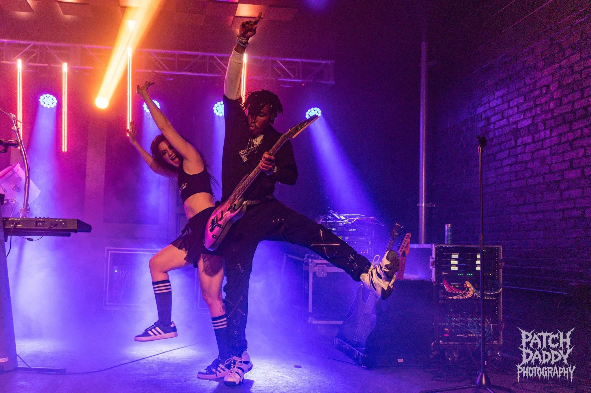 A male guitarist and a female dancer perform on stage with vibrant lighting. The guitarist is in the foreground, playing an electric guitar, with his leg raised. The female dancer is behind him, dancing with her arms raised. The stage has purple and 