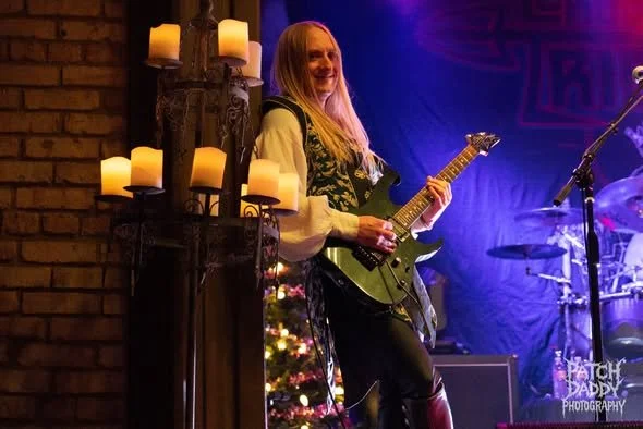 A woman playing an electric guitar on stage with colorful lighting, candles, and a small decorated Christmas tree nearby.
