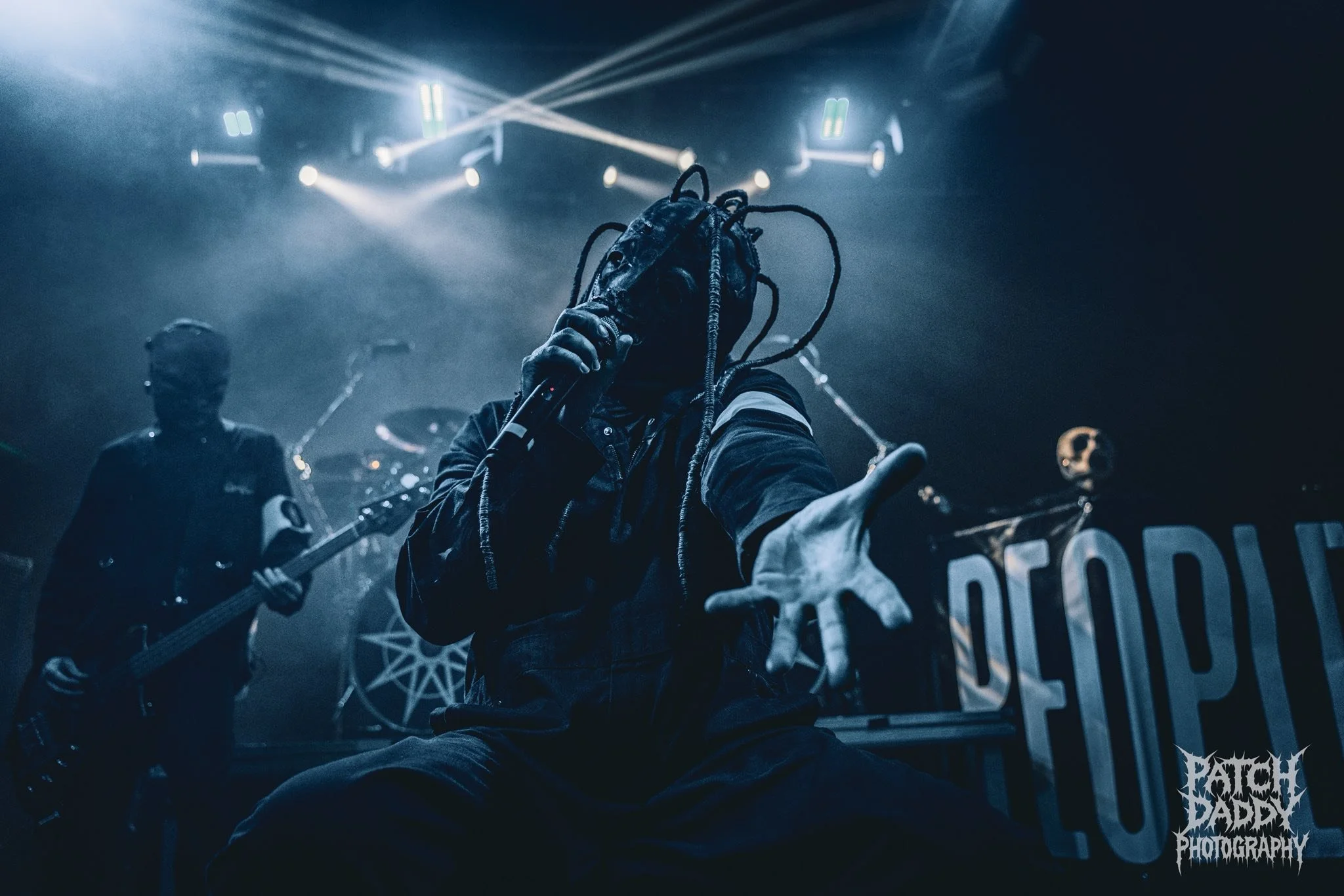 A performer with dreadlocks and dark clothing holding a microphone on stage, with band members playing guitars in the background, under blue lighting.