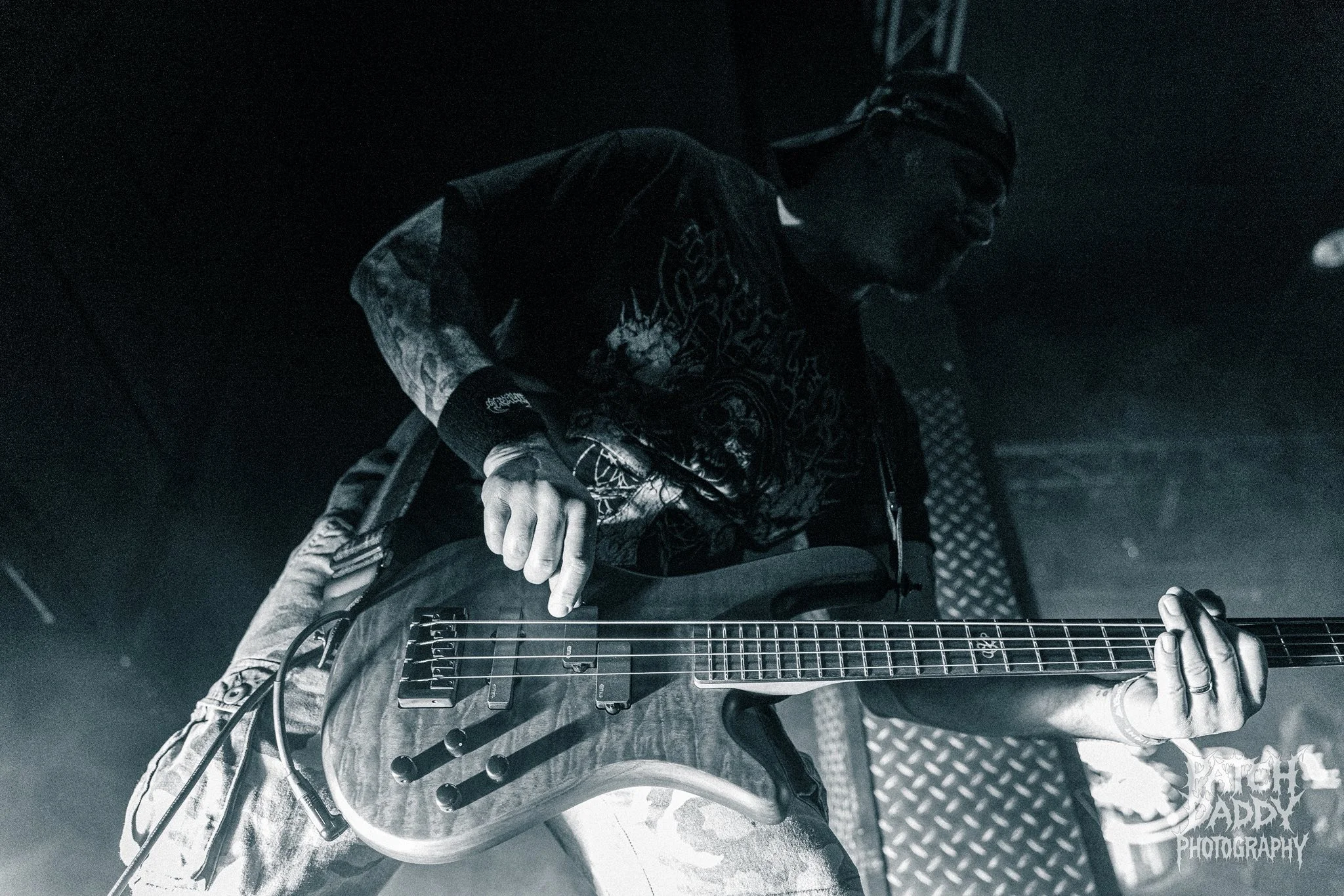 A musician playing an electric guitar on stage in black and white, wearing glasses, a cap, a band t-shirt, and camouflage pants.