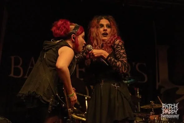 Two women with vibrant hair, one with pink and the other with red, on stage during a musical performance, one holding a microphone.