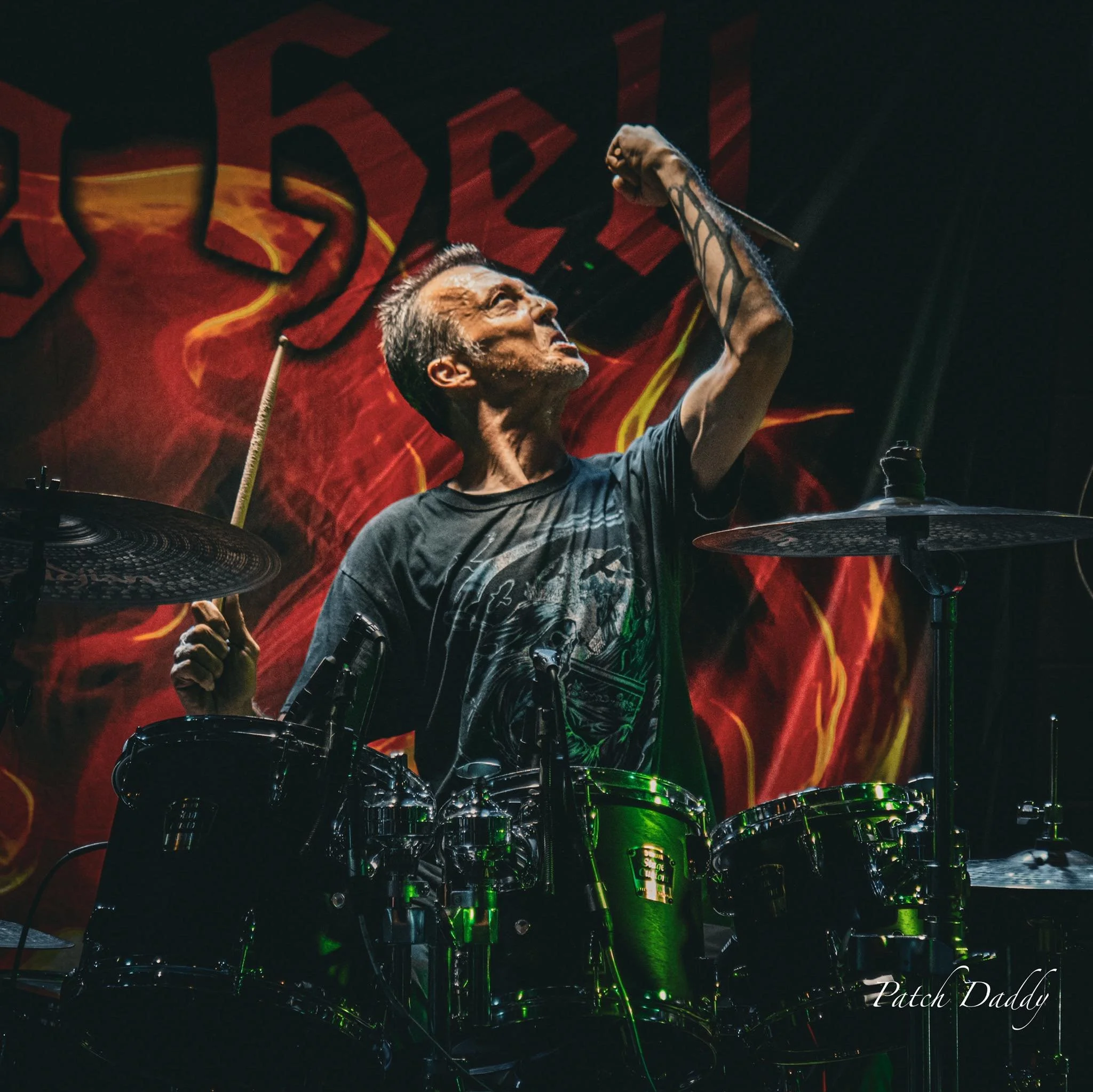 A drummer on stage with drum kit, raising a drumstick in the air, with a smoky, fiery backdrop.