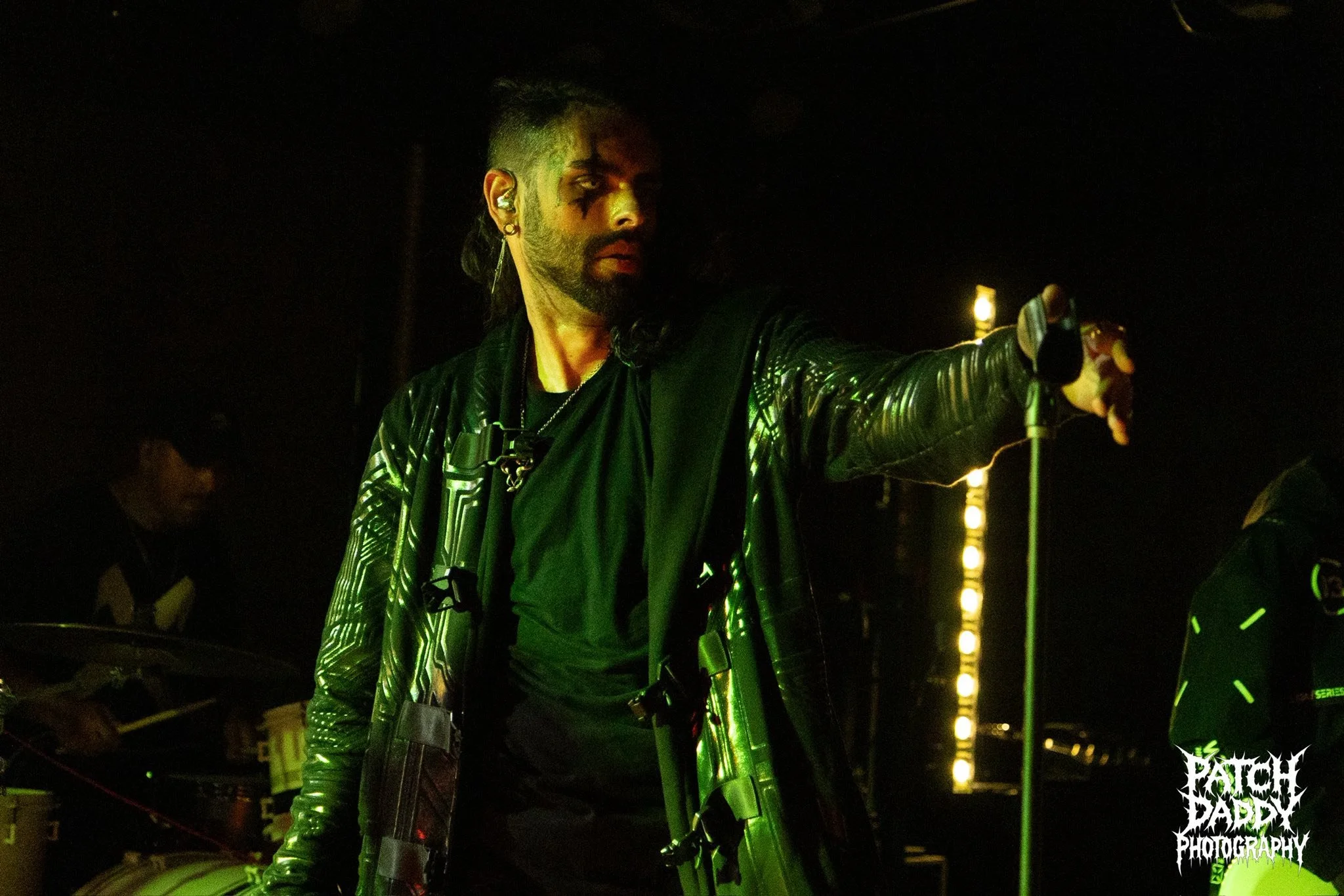A person with dark hair and makeup, wearing a shiny black jacket, performing on stage with a microphone in hand, illuminated by green lighting.