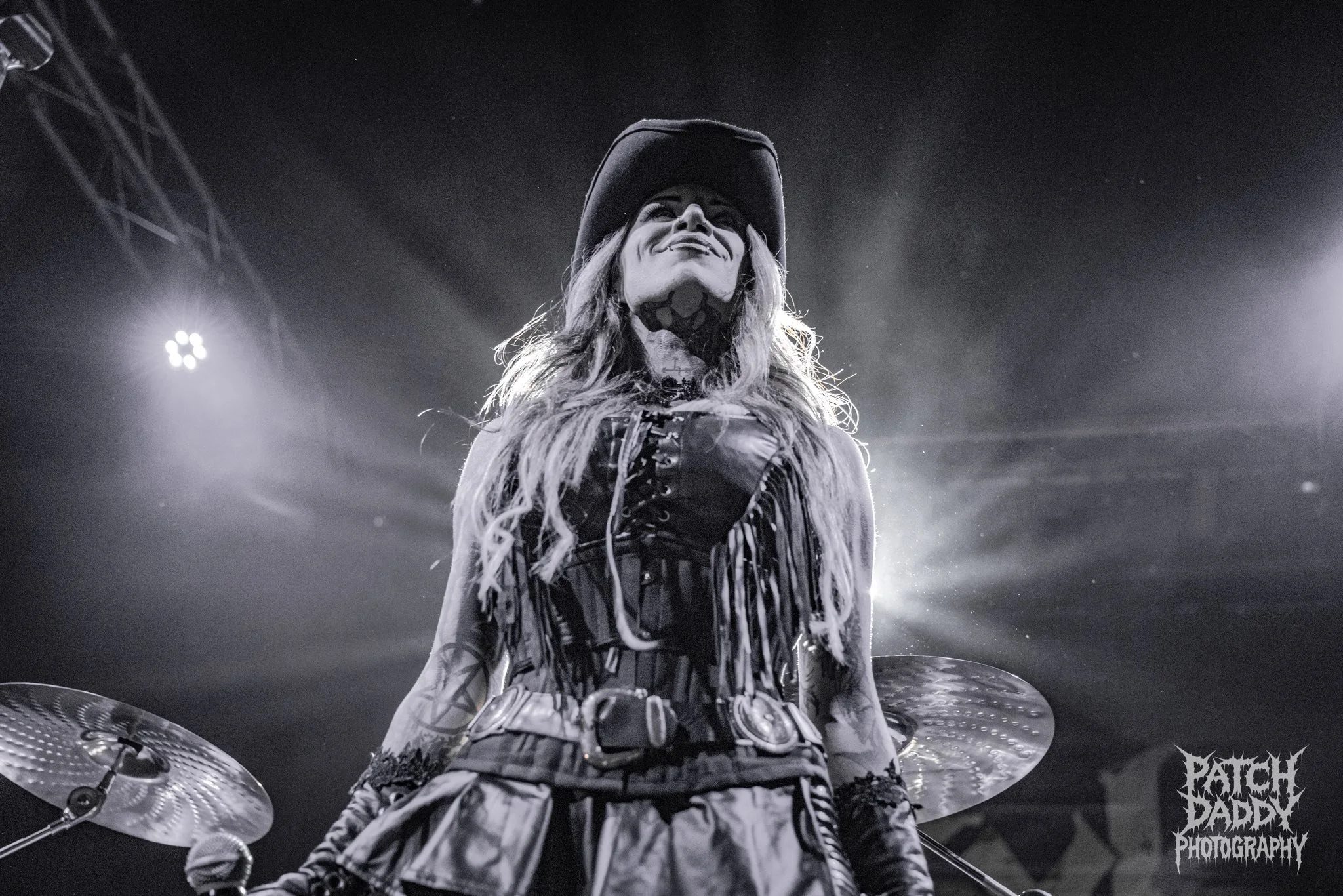 A woman dressed in a pirate costume on stage, smiling with long wavy hair, wearing a hat, corset, and accessories, with drums and stage lights in the background.