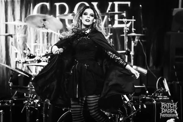 A woman in gothic-style clothing performing on stage with a microphone, with drums in the background and the word 'CROWE' visible on a banner.