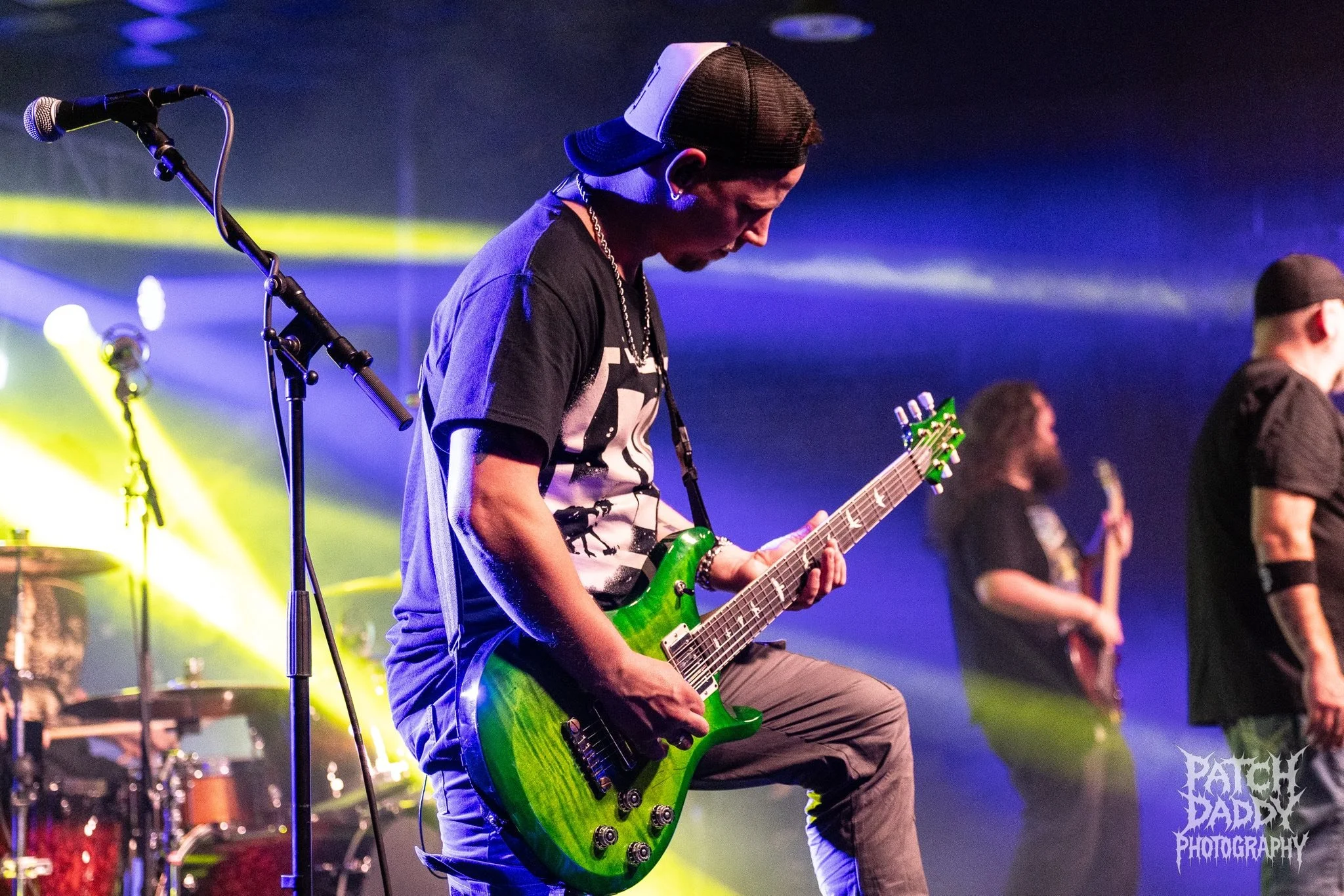 A male guitarist in a black cap, black t-shirt, and grey pants playing a green electric guitar on stage with yellow and purple stage lights.