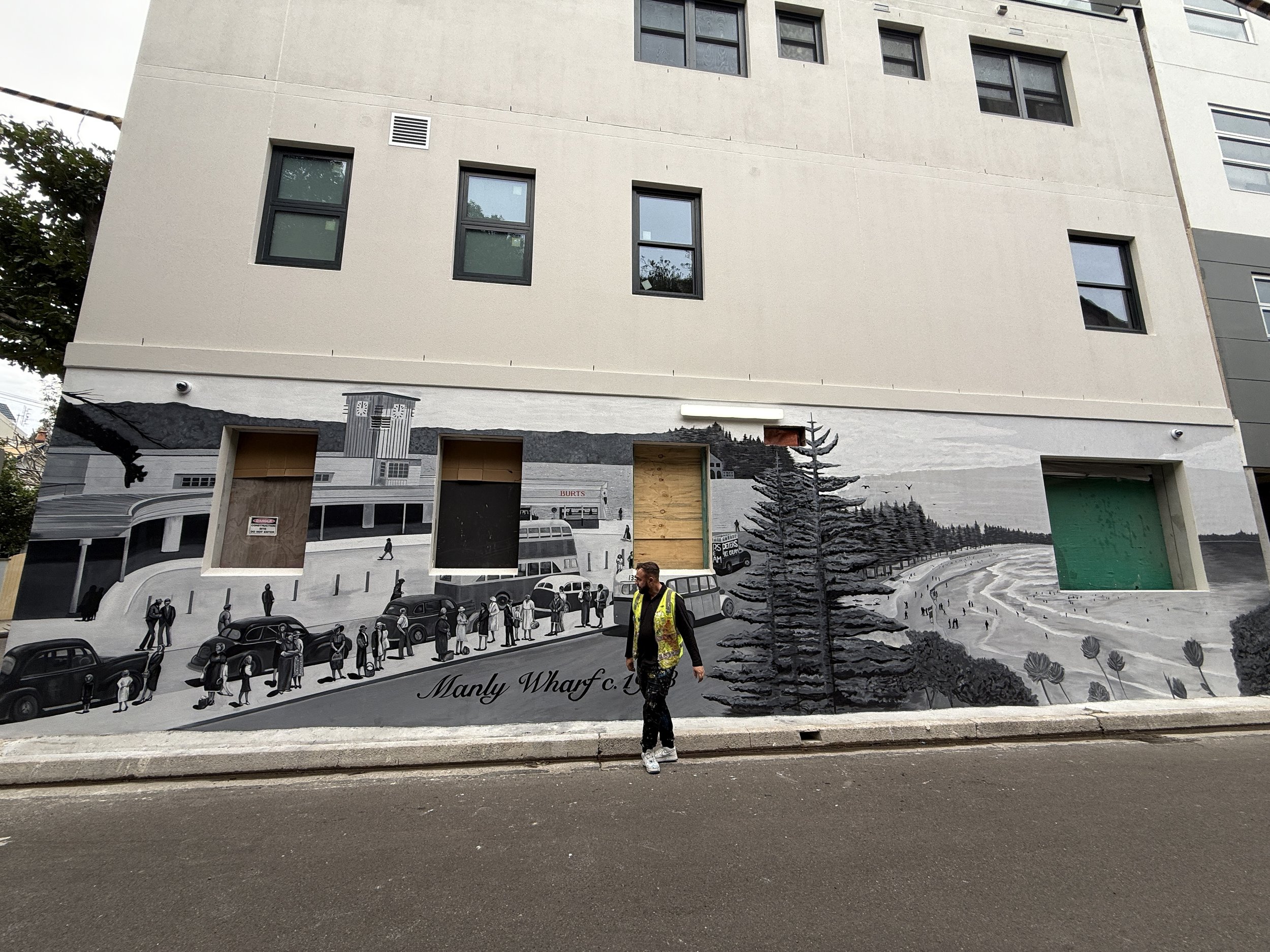Mural depicting a black-and-white city scene with cars, pedestrians, a bus, trees, and a shoreline, painted on the lower part of a beige building's wall, with a man in a yellow vest walking in front. Mural of Manly Wharf C.1943, Hand painted