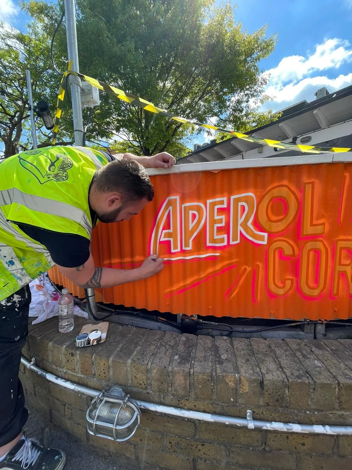 A man in a fluorescent yellow safety vest is painting a sign that says "APEROL CORNER" with a neon pink and orange outline, while standing on a brick sidewalk under a partly cloudy sky. Hand Painted Signwriting for Aperol Activation