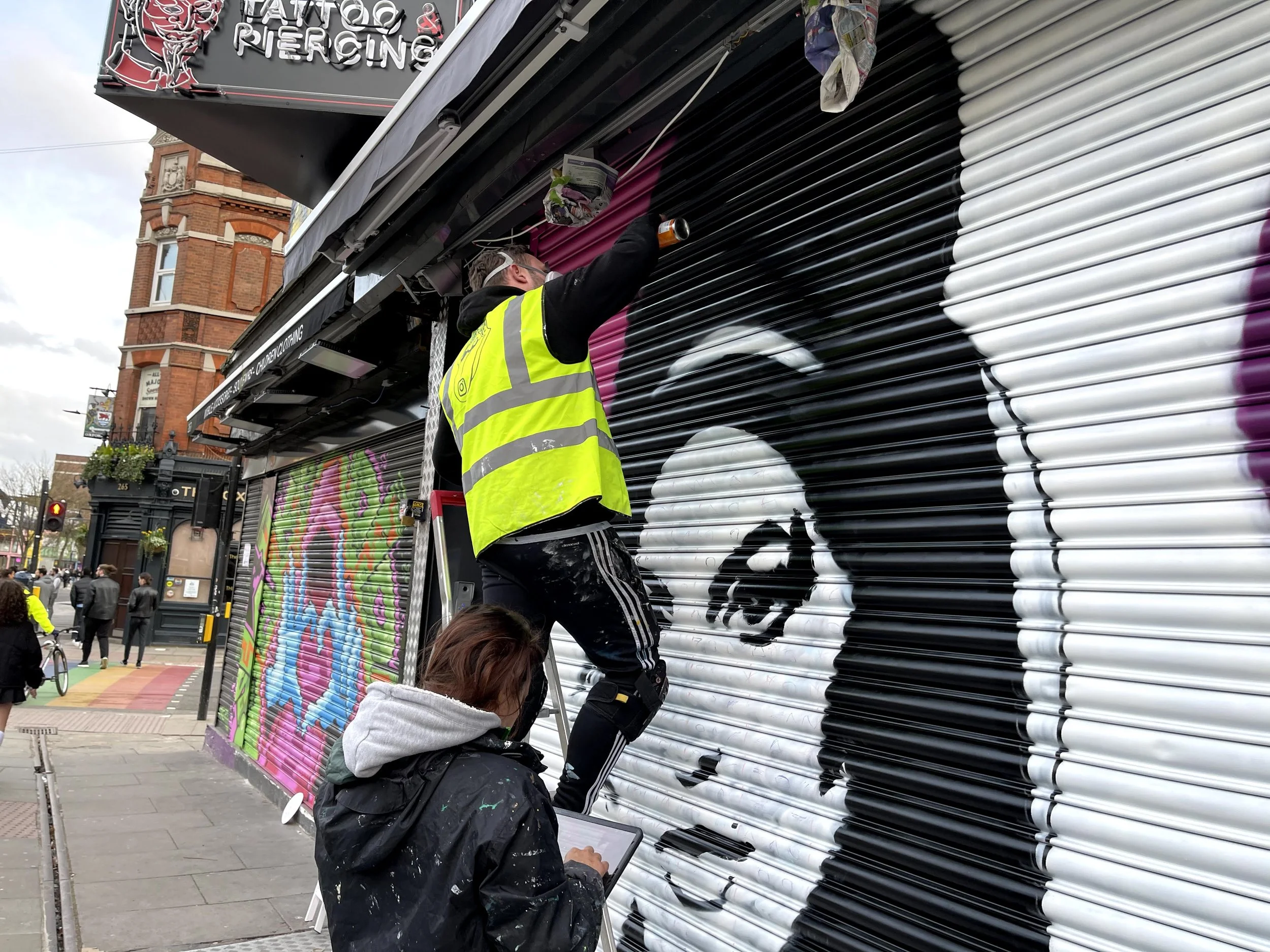 Two artists are painting a mural of a woman's face on a metal roller shutter in an urban street. One artist is standing on a step ladder, applying paint, while the other is crouching and working on a tablet. The mural features a monochrome portrait of a woman with dark hair and makeup.