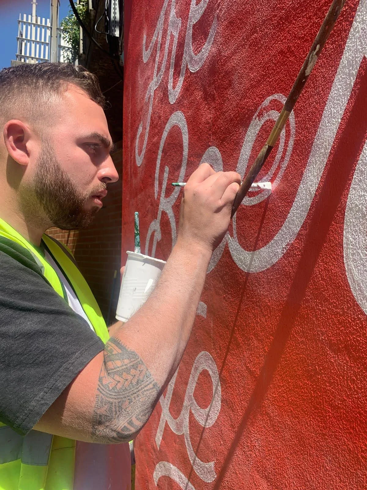 A man with a beard and a tattoo on his arm is painting white text on a red wall using a thin brush, holding a small cup of white paint.