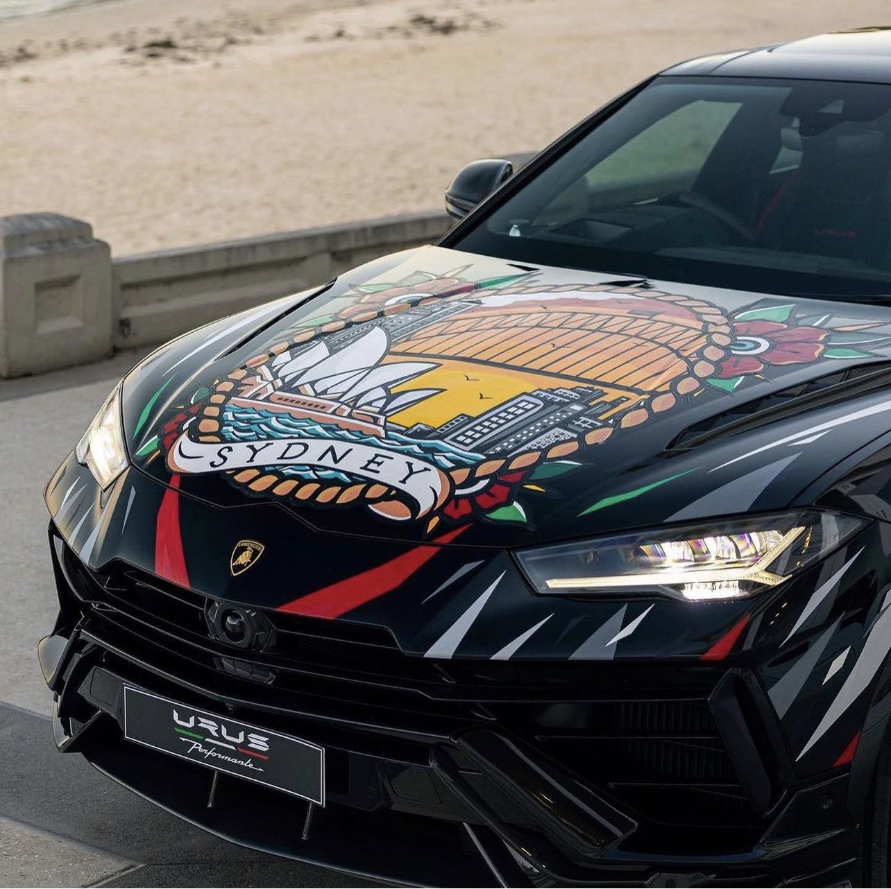 A black Lamborghini with a colorful graphic of a Sydney-themed design on the hood, including the Sydney Opera House, Harbour Bridge, and cityscape. Live Painted Lamborghini Graffiti Tattoo art style