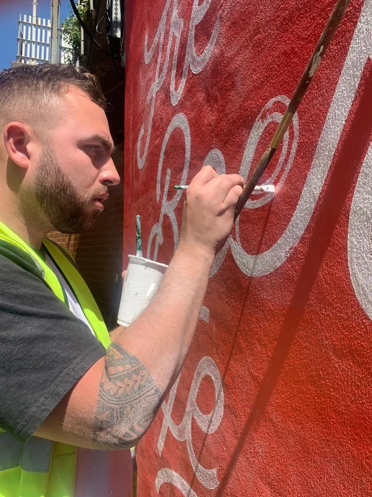 A man with a beard and a tattoo on his arm is painting white lettering on a red wall. He is holding a paintbrush in one hand and a container of white paint in the other. The man is wearing a black shirt and a yellow safety vest.