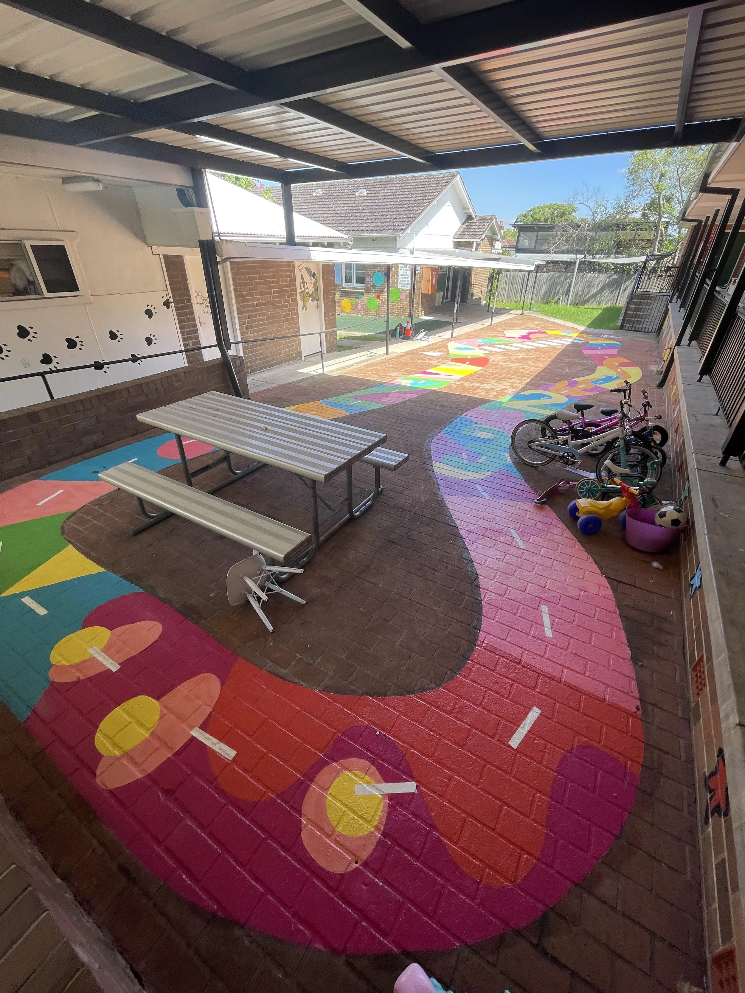 Colorful outdoor play area with a rainbow winding path painted on brick ground, surrounded by toys, bicycles, picnic tables, and a covered patio at a school or daycare.