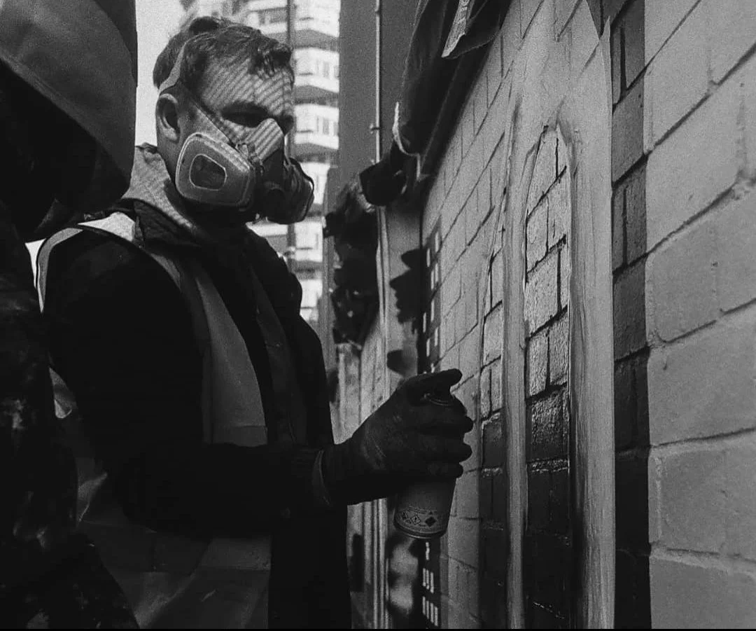 A person wearing a respirator mask, gloves, and a reflective safety vest is using a spray paint can to paint graffiti on a brick wall in SYDNEY