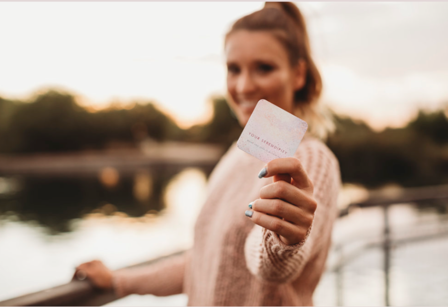 A woman with reddish hair wearing a beige sweater is holding a small card toward the camera, with a blurred outdoor background possibly near a body of water during sunset.