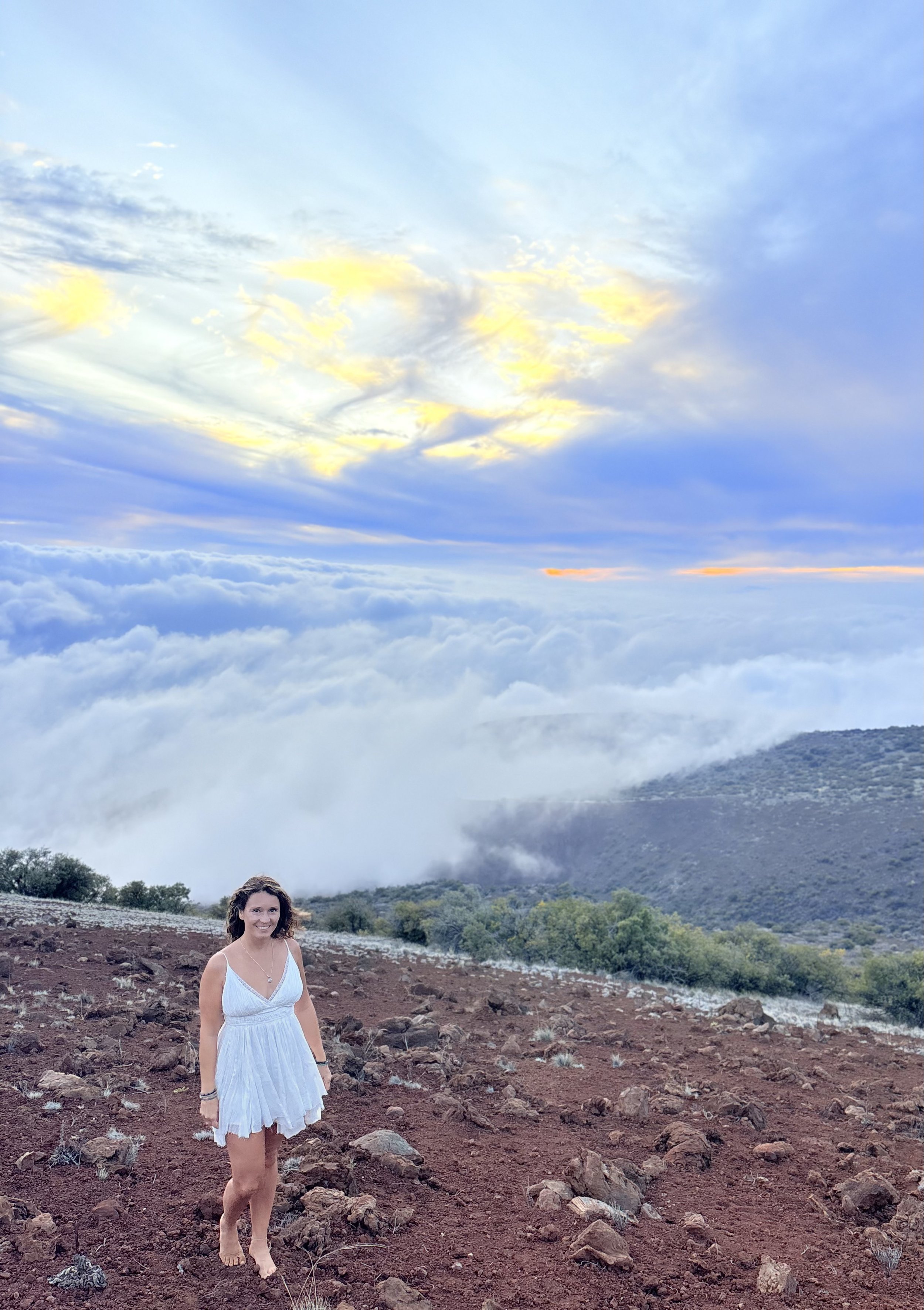 A woman in a white dress standing on rocky terrain with a mountain and cloudy sky in the background.