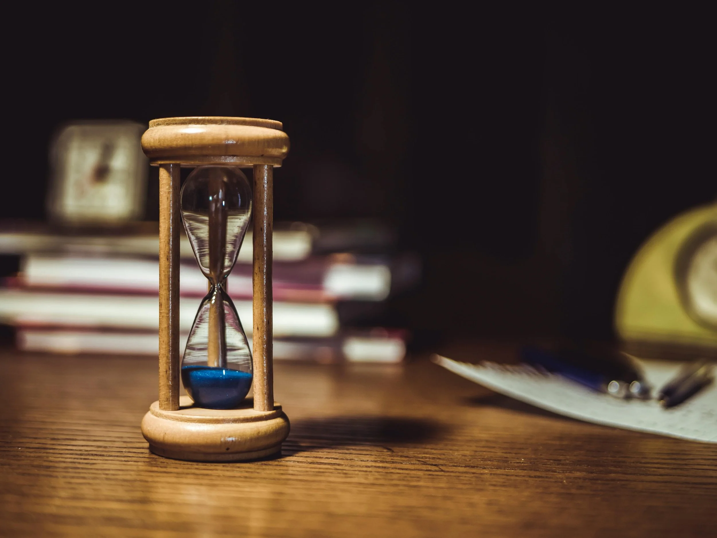 Small wooden hourglass with blue sand on a wooden desk, blurred notebooks and a clock in the background.