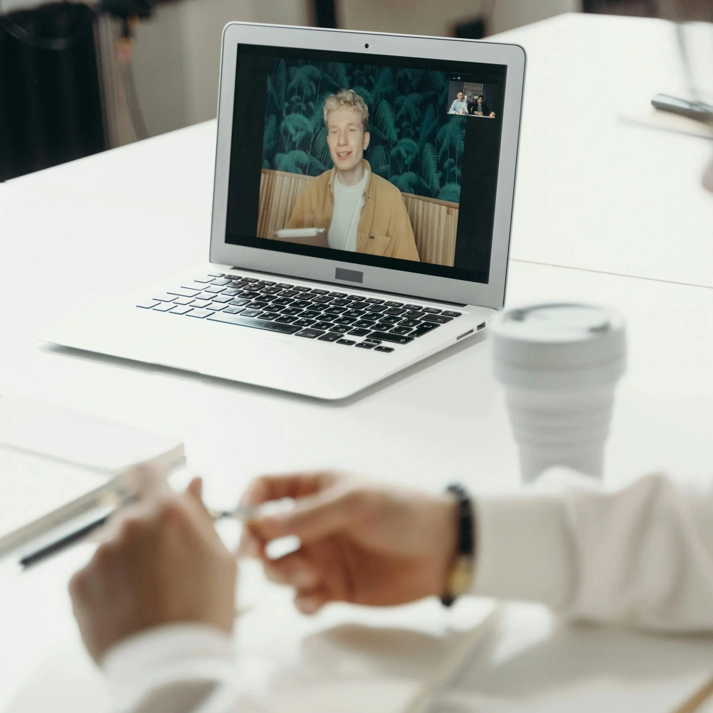 A laptop on a white table displaying a video call with a young man speaking. Someone's hands are visible in the foreground, taking notes in a notebook with a pen. A disposable coffee cup is also on the table.