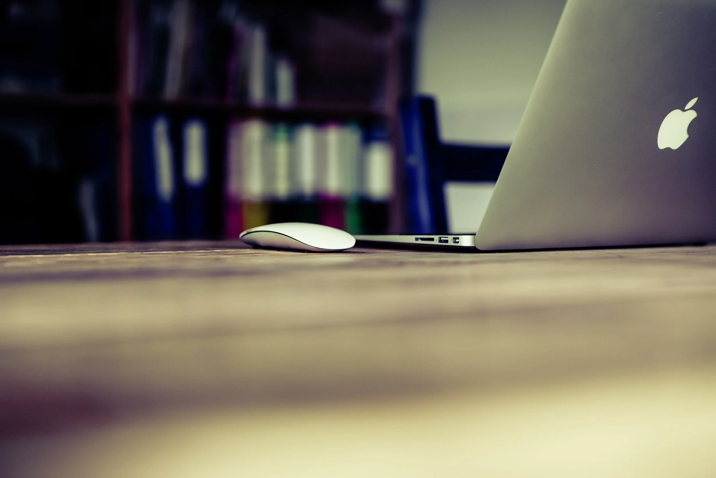 Close-up of a white Apple MacBook laptop and a white wireless mouse on a wooden desk, with a blurred background of a bookshelf filled with books.